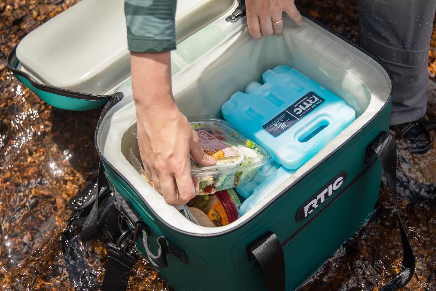 Man grabbing snack out of RTIC soft cooler bag that is sitting in river