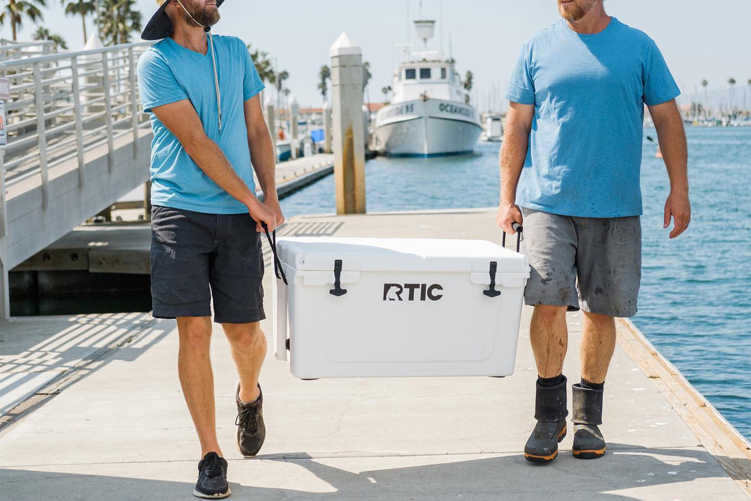 Two men carrying white RTIC 110 quart cooler on a boat dock.