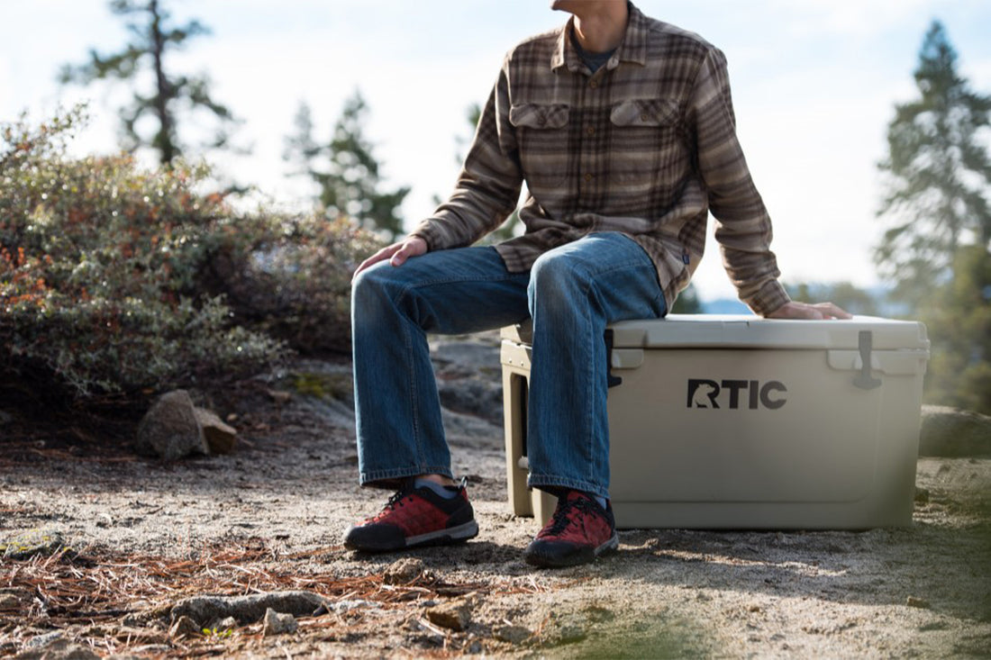 Man sitting on gray RTIC 65 quart cooler in the woods.