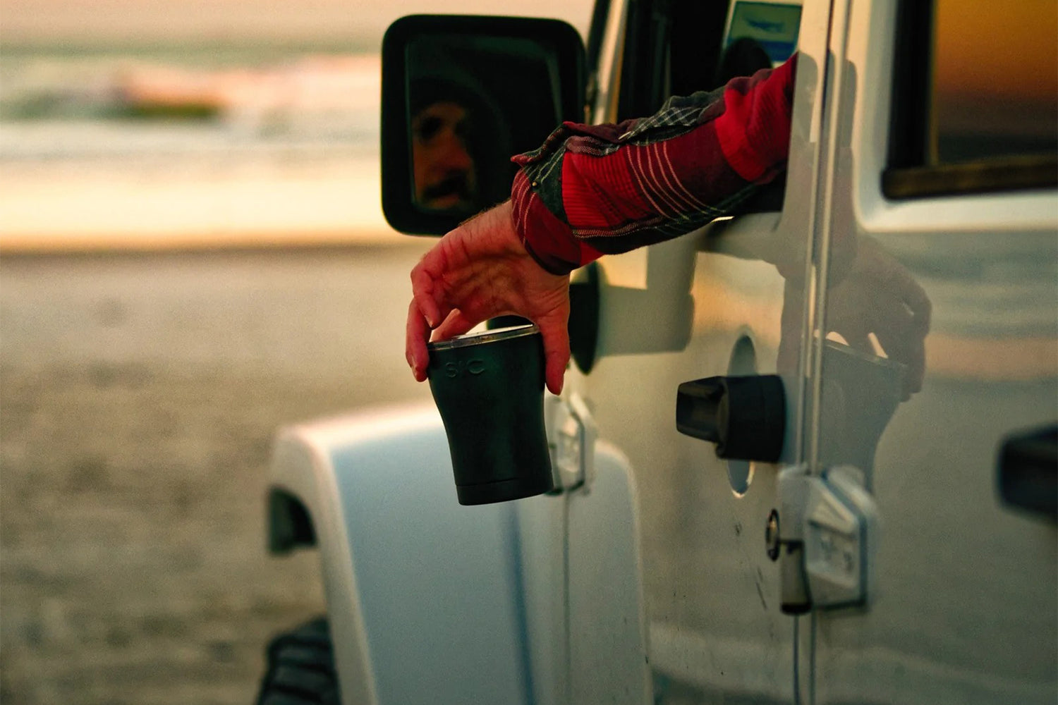 Man holding black SIC tumbler sitting in jeep on the beach at sunset