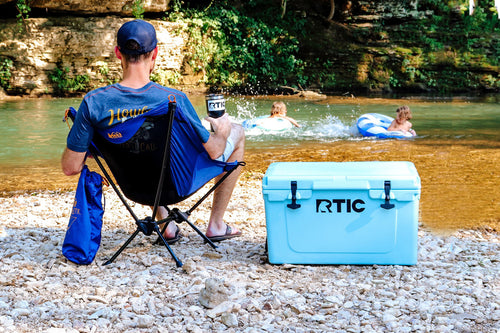 Man sitting at swimming hole while kids swim. Blue RTIC 45 quart hard cooler sitting on ground.