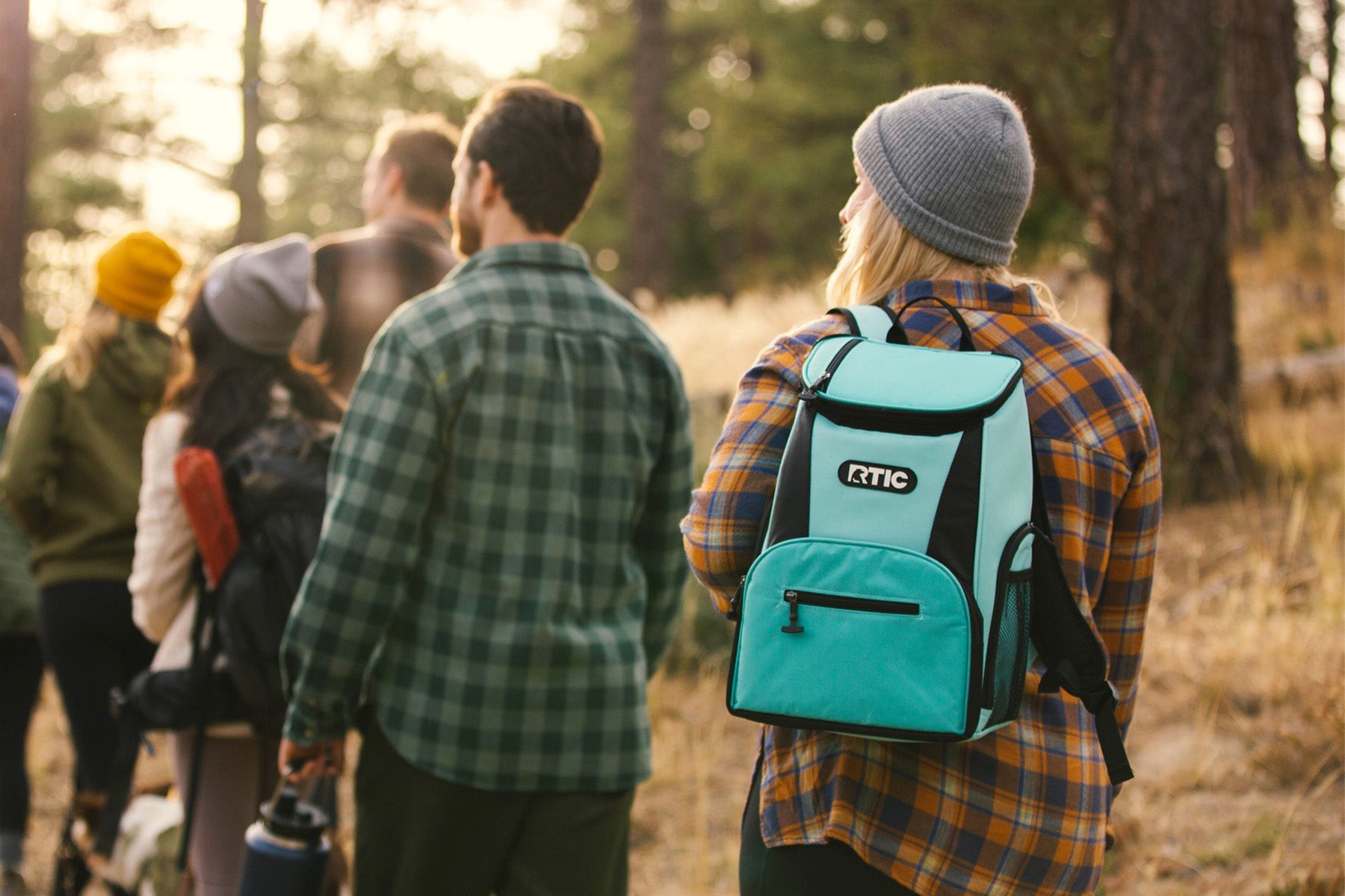 Group of people hiking through woods. Woman has RTIC backpack cooler in Aqua color