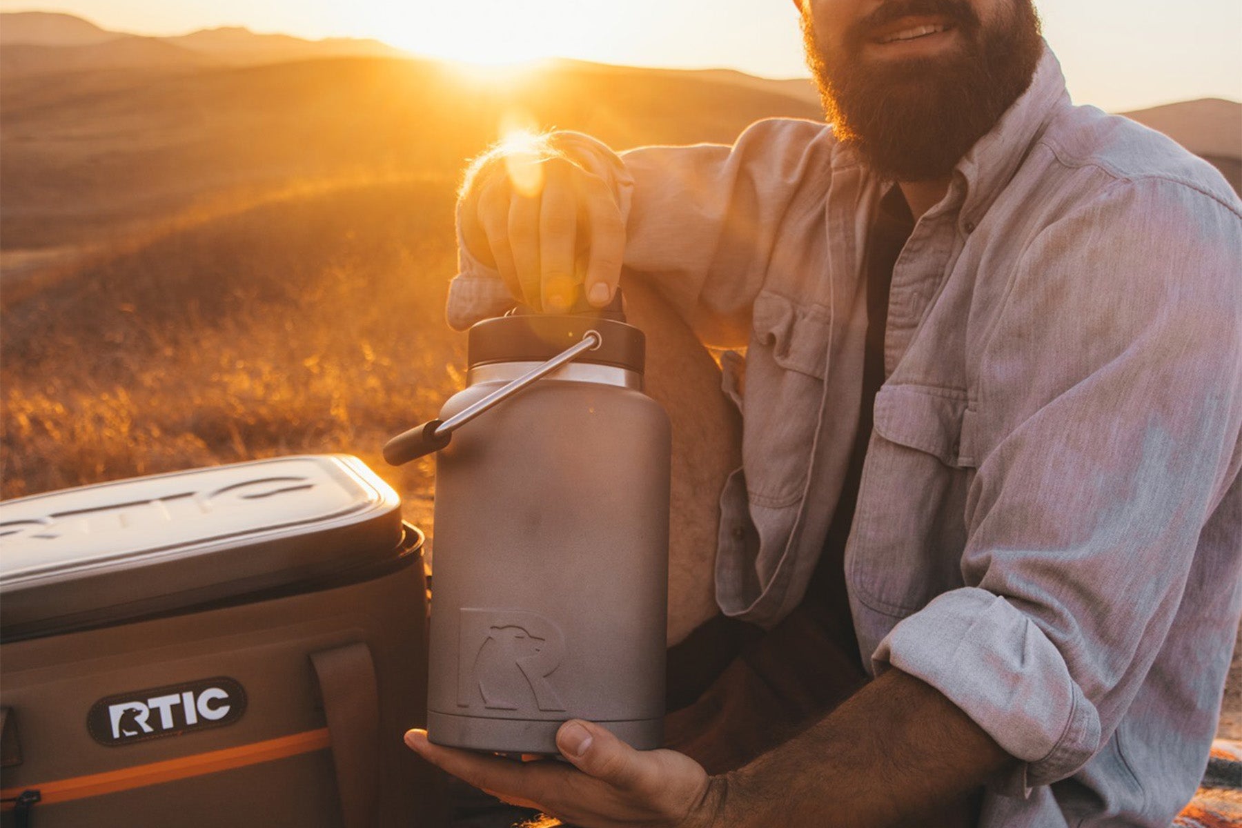 Man holding gray RTIC Half Gallon water jug next to tan RTIC cooler outside