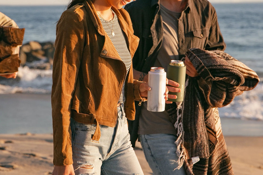 Friends walking on a beach while holding Brumate Hopsulator Slim can holder koozies in white and green
