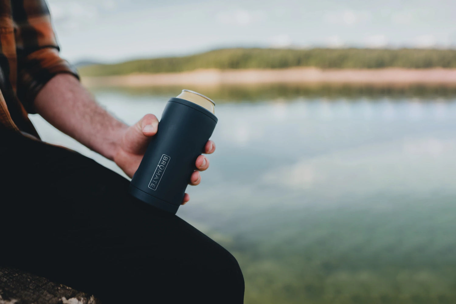 Man sitting outside by a lake holding a black Brumate Hopsulator Trio can holder koozie